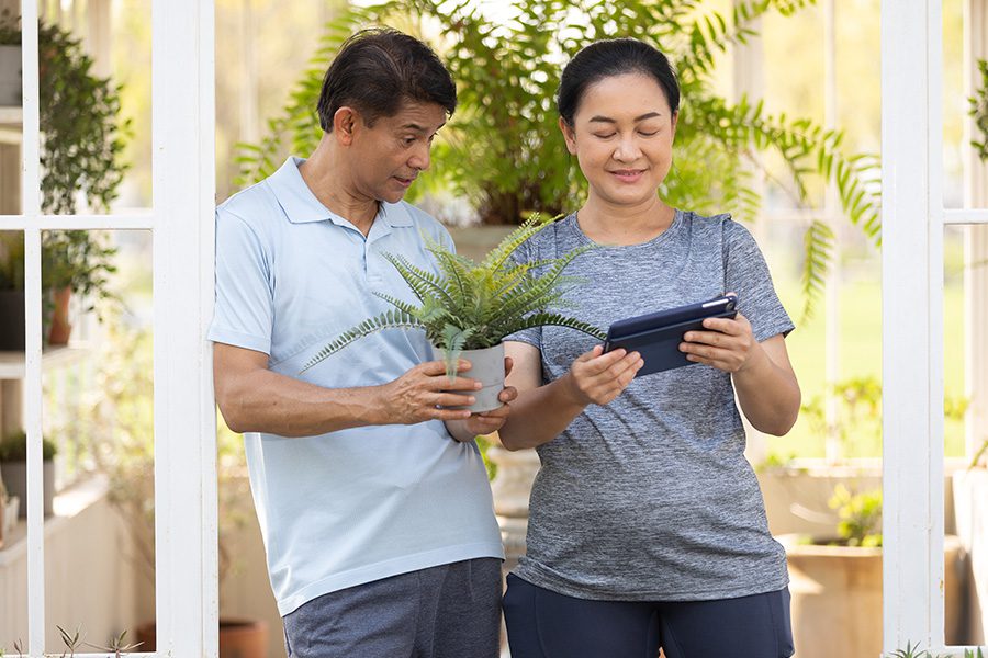 Client Center - Senior Couple Smiling While Holding Plant Pots and Using a Tablet in Their Garden